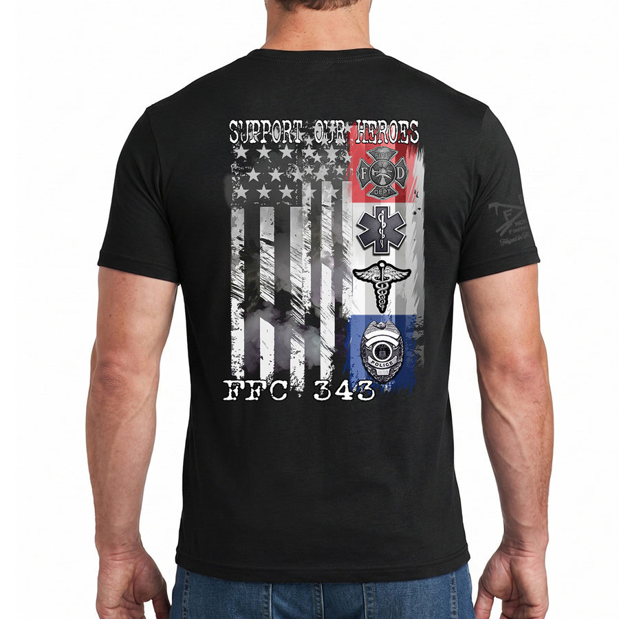 A man wearing a black t-shirt, viewed from the back. The shirt features a large graphic print on the back with a distressed American flag, a red fire department cross, a medical emergency cross, and a blue first responder helmet, with the text 'SUPPORT OUR HEROES' and 'FFC 343'.