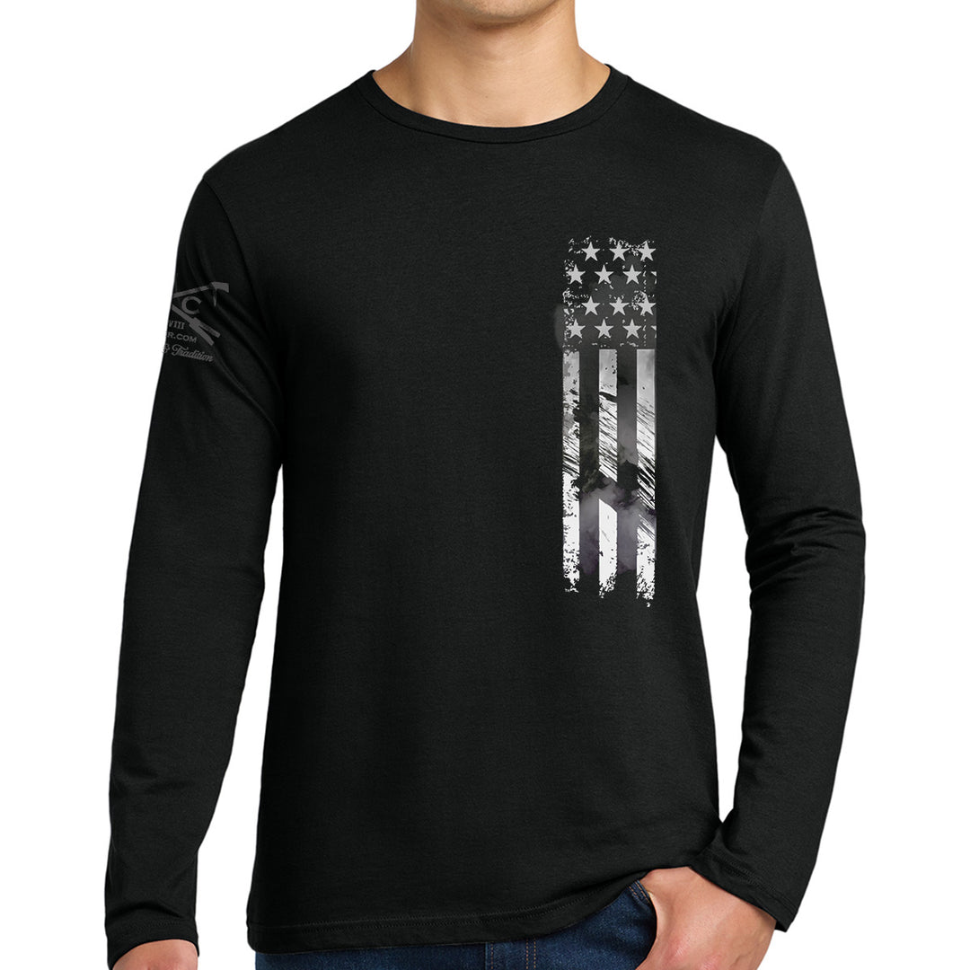 A man wearing a black long-sleeve t-shirt with a distressed vertical American flag graphic in white and gray on the left chest, shown against a plain white background. #color_black