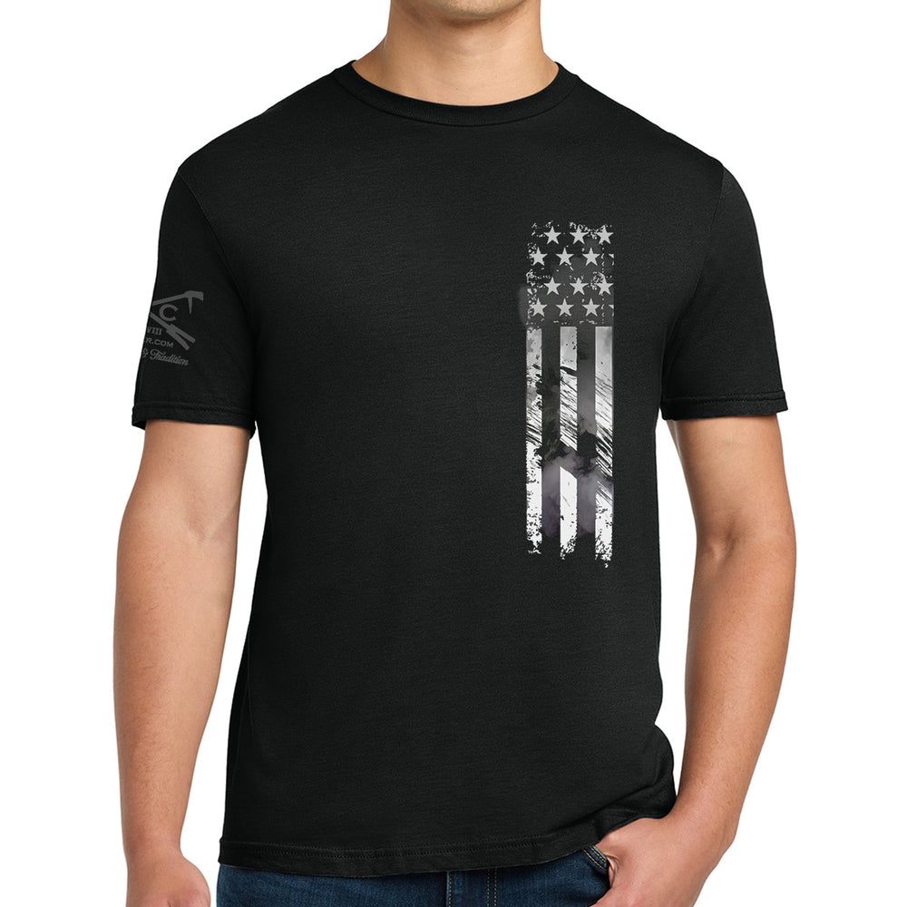 A man wearing a black short-sleeve t-shirt with a distressed vertical American flag graphic in white and gray on the left chest, and a small dark graphic on the right sleeve.