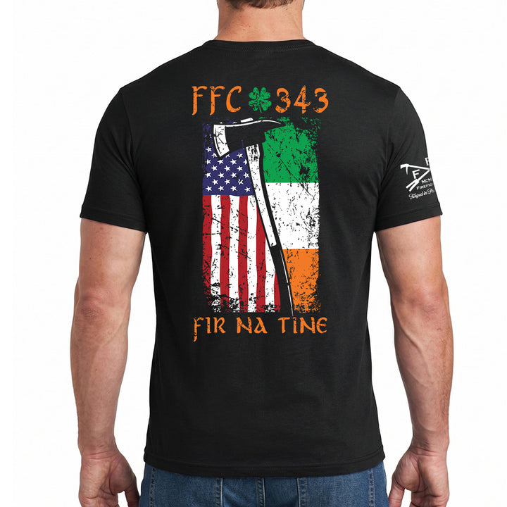 A man wearing a black t-shirt, viewed from the back. The shirt features a large graphic print on the back with the text 'FFC 343' and 'FIR NA TINE' in orange, a shamrock, and a distressed vertical split flag showing the American flag on the left and the Irish flag on the right. #color_black
