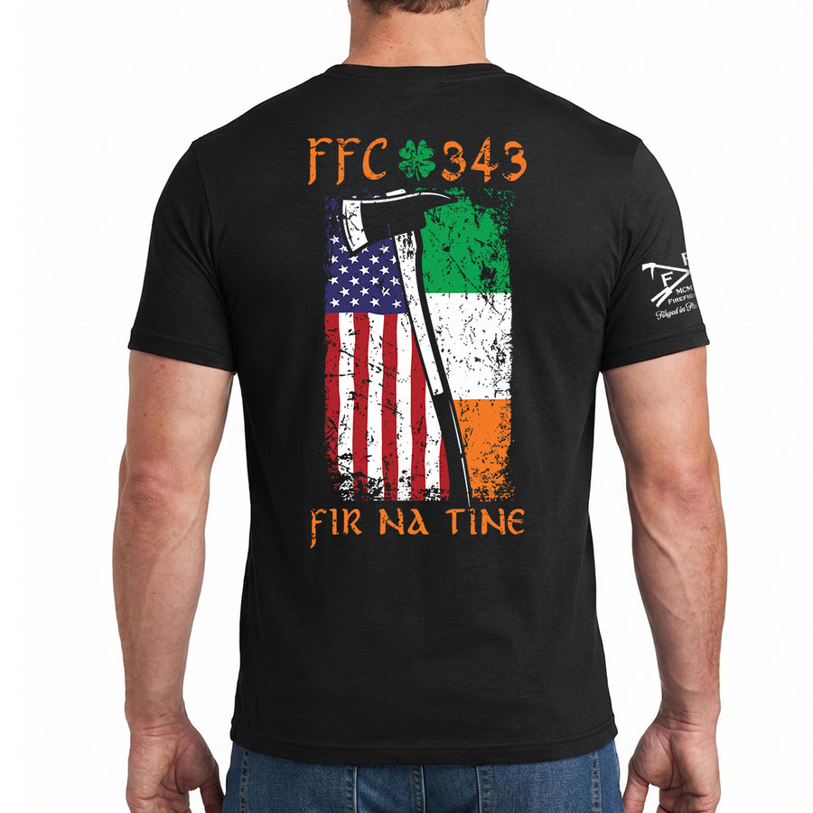 A man wearing a black t-shirt, viewed from the back. The shirt features a large graphic print on the back with the text 'FFC 343' and 'FIR NA TINE' in orange, a shamrock, and a distressed vertical split flag showing the American flag on the left and the Irish flag on the right. #color_black