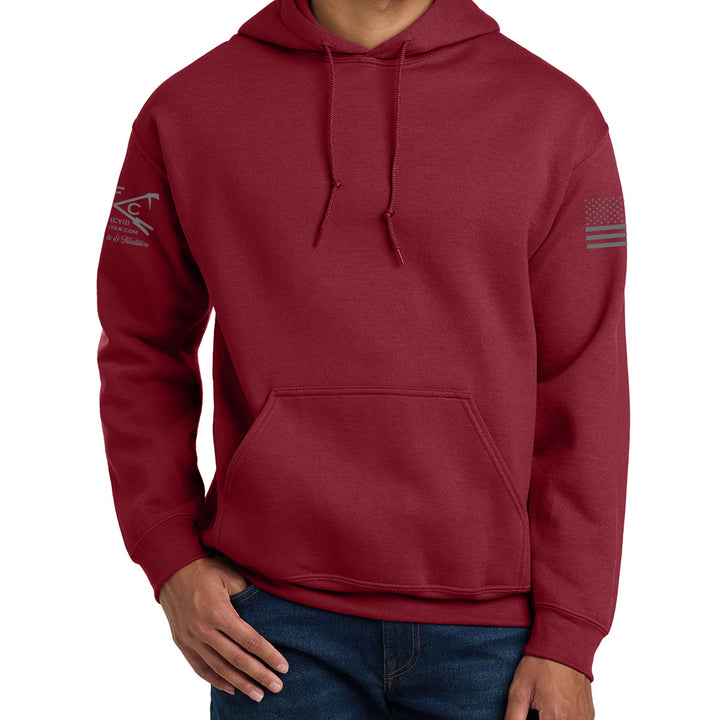A man wearing a maroon pullover hoodie with a front kangaroo pocket and drawstrings. The hoodie features a dark gray graphic logo on the left sleeve and a dark gray American flag graphic on the right sleeve. #color_cardinal-red