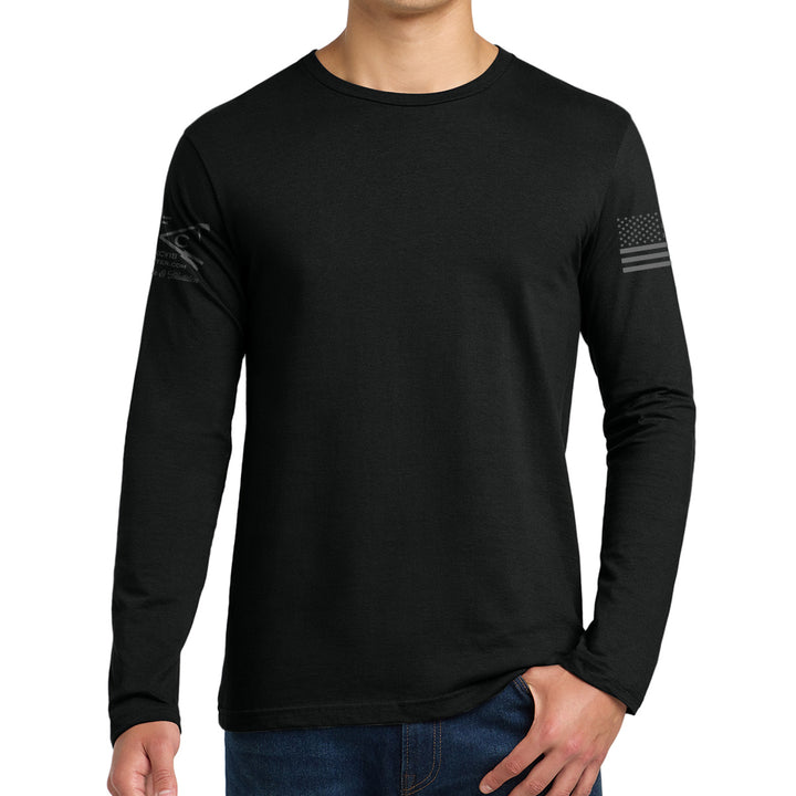 A man wearing a black long-sleeve crewneck shirt with a dark gray distressed American flag graphic on the left sleeve, paired with blue jeans against a plain white background. #color_black