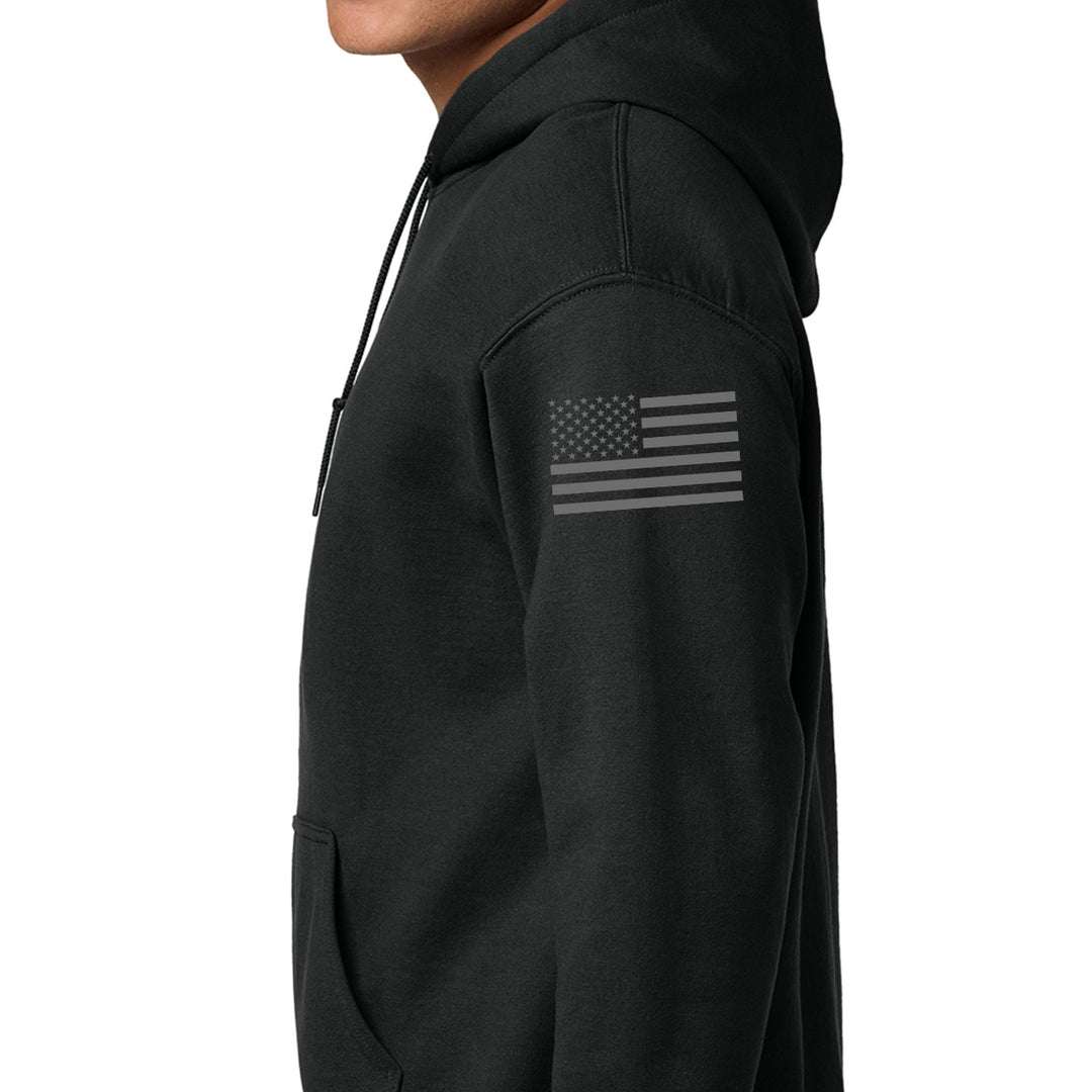 A close-up side view of a person wearing a black pullover hoodie with a dark gray distressed American flag graphic printed on the upper left sleeve, set against a plain white background. #color_black