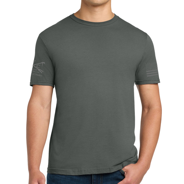 A man wearing a dark gray crewneck t-shirt with a subtle tonal graphic on the left sleeve and an American flag print on the right sleeve, paired with blue jeans against a white background. #color_charcoal