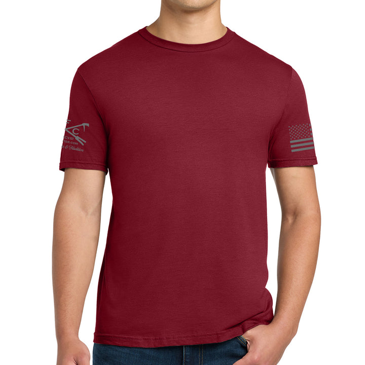 A man wearing a short-sleeve crewneck t-shirt in maroon red, featuring a gray tactical logo on the left sleeve and a distressed American flag graphic on the right sleeve, shown against a plain white background. #color_cardinal-red
