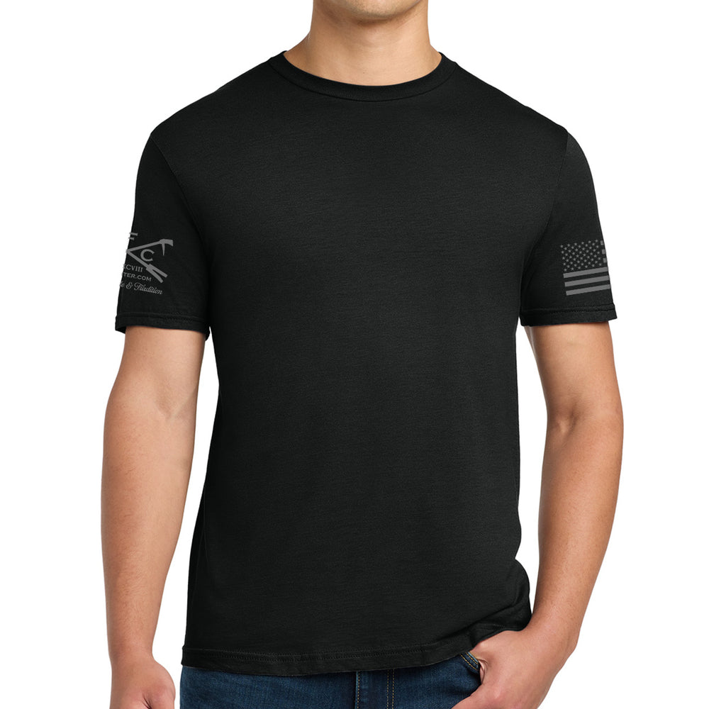 A man wearing a black short-sleeve crewneck t-shirt with a dark gray distressed American flag graphic on the left sleeve and a tactical cross graphic on the right sleeve, paired with blue jeans against a white background. #color_black