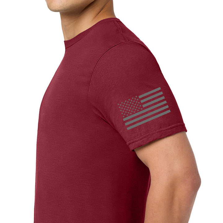 A man wearing a burgundy short-sleeve t-shirt with a dark gray distressed American flag graphic printed on the left sleeve, shown against a plain white background. #color_cardinal-red