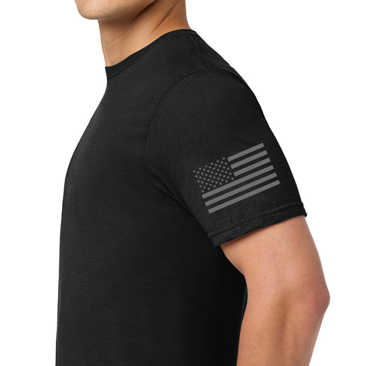A man wearing a black short-sleeve t-shirt with a subtle, distressed gray American flag graphic printed on the left sleeve, shown against a plain white background. #color_black