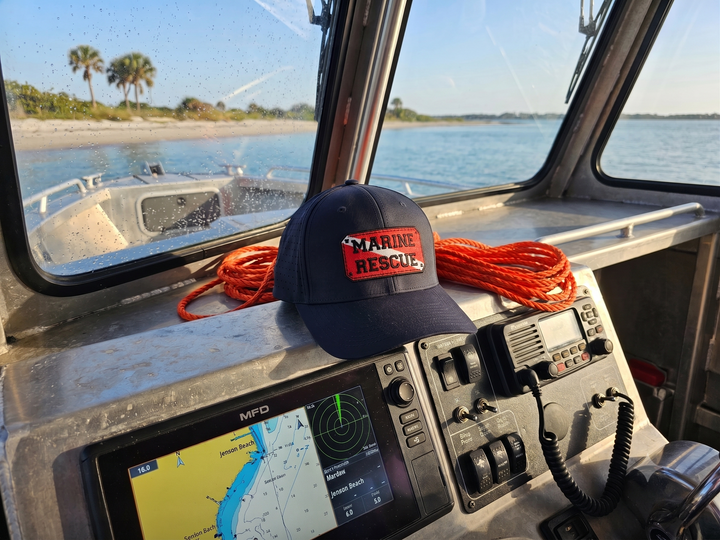 A navy blue baseball cap with a red and white 'MARINE RESCUE' patch on the front, resting on the dashboard of a boat cabin alongside coiled orange ropes and marine electronics.